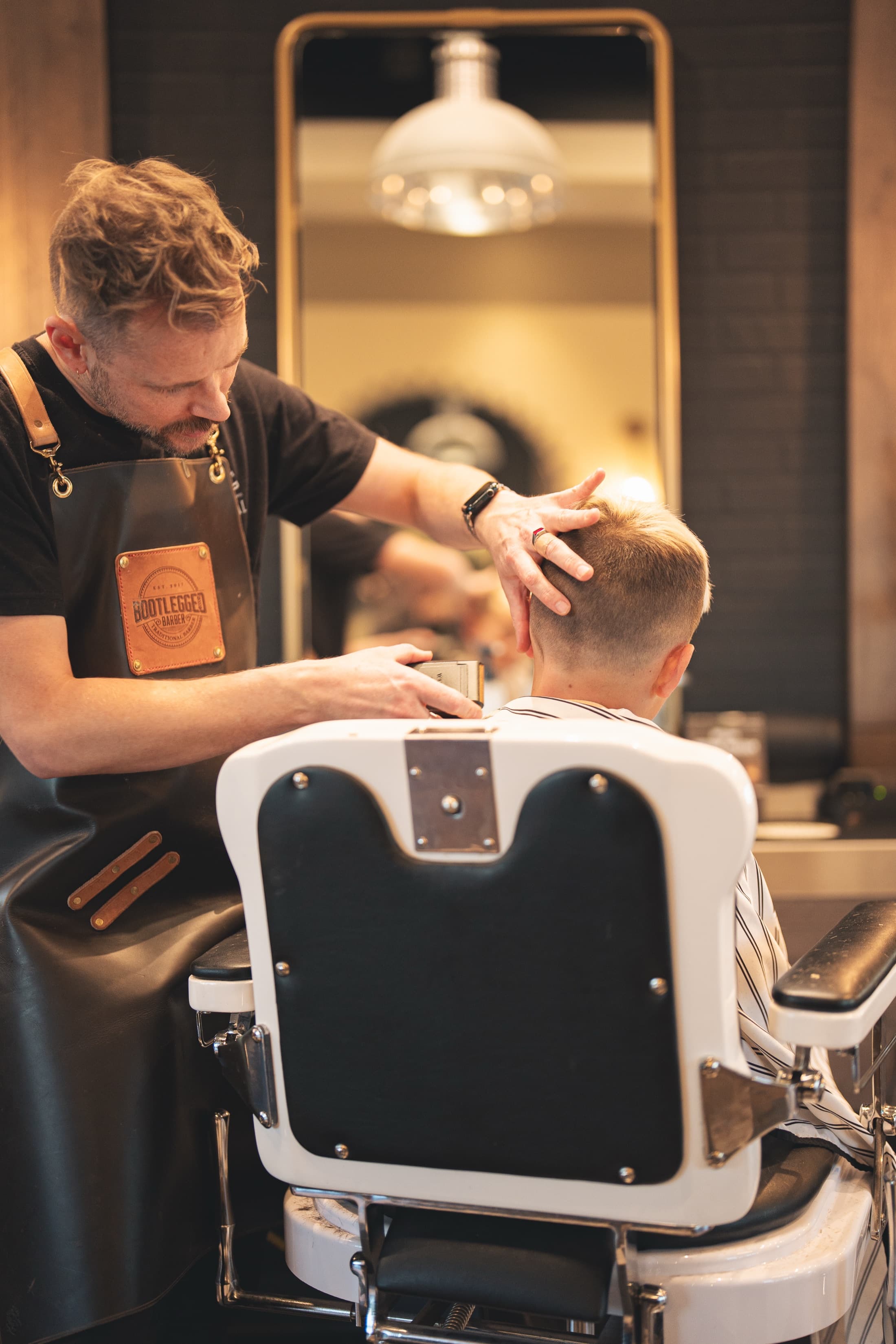 Barber in a leather Bootlegged apron guiding a clipper up a young client's fade